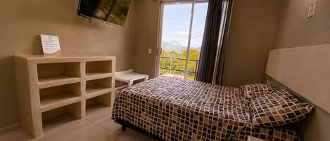 A bedroom with a bed, a TV mounted on the wall, a window with curtains, and a small table.