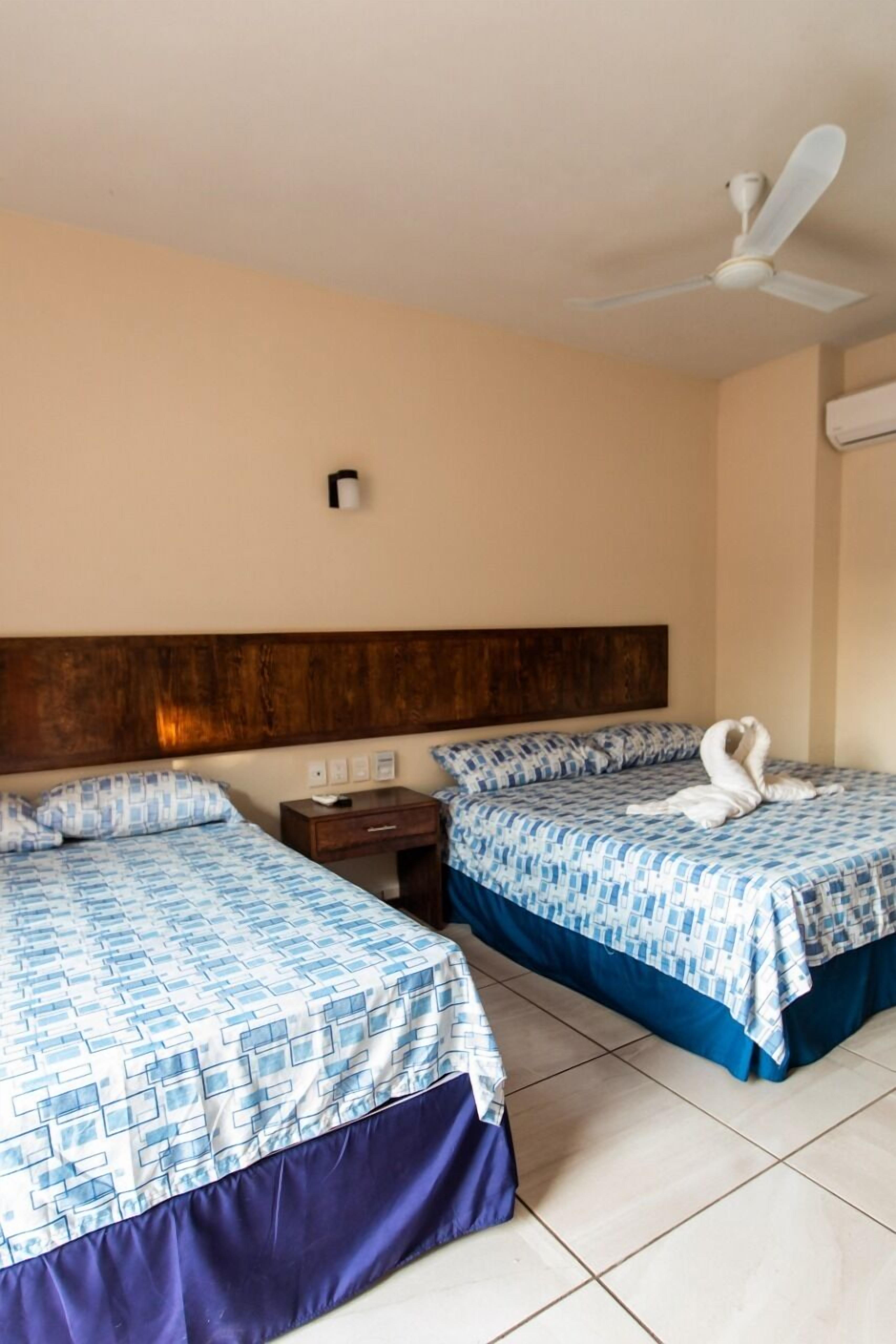 Une chambre d’hôtel avec deux lits, une tête de lit en bois, un ventilateur de plafond et une table de chevet avec une lampe.