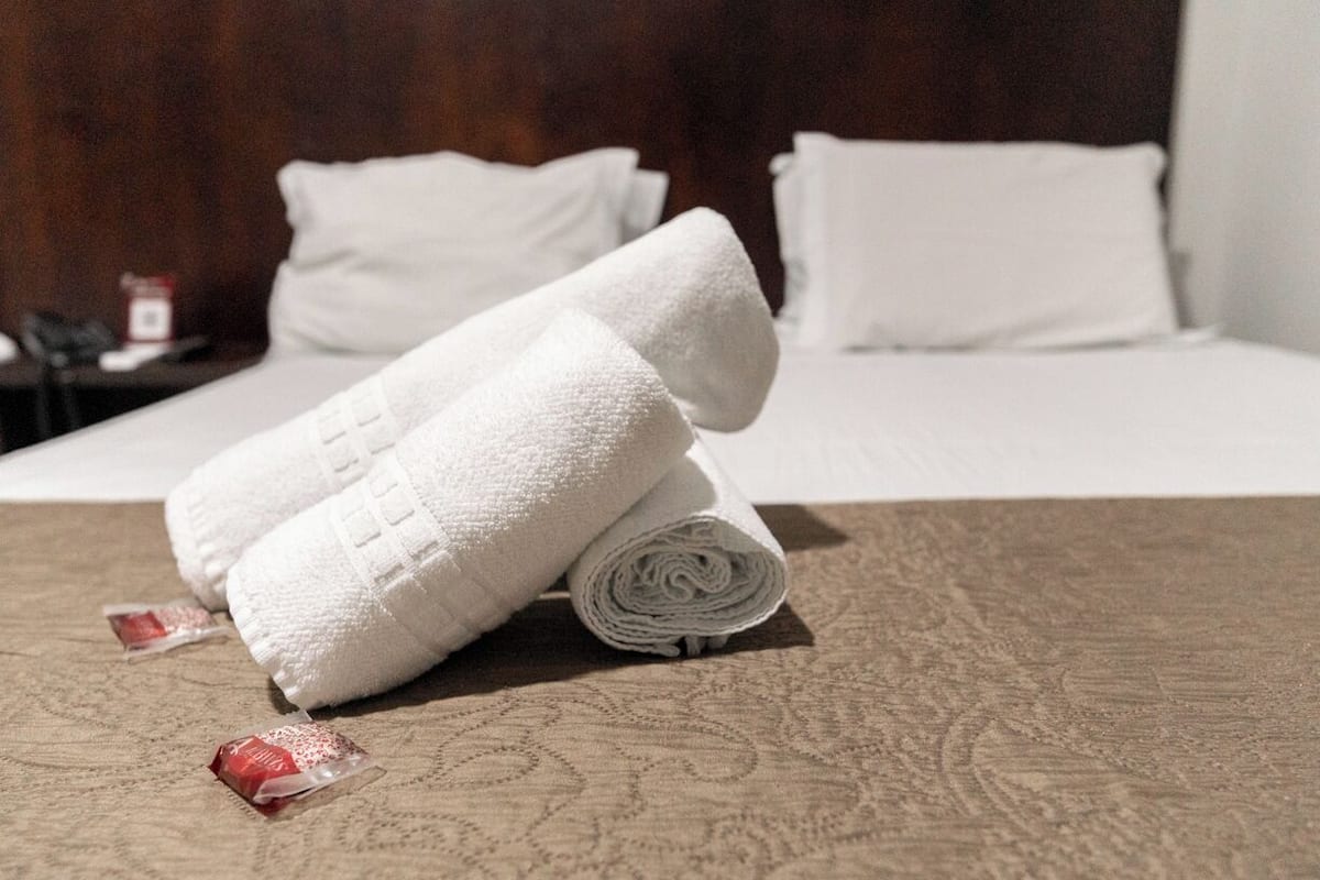 Cama bem arrumada com roupa de cama branca e uma toalha enrolada sobre um edredom texturizado.