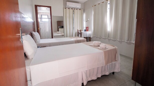 A hotel room with two beds, a bathroom, and a small table with a chair. - Hotel Albinos (Itaperuna)