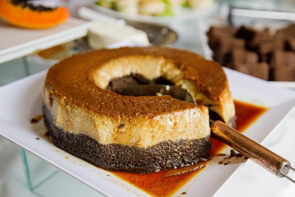 Um bolo de sobremesa sobre um prato branco, com cobertura caramelizada e base escura, possivelmente de chocolate, acompanhado de molho de caramelo e um pau de canela.