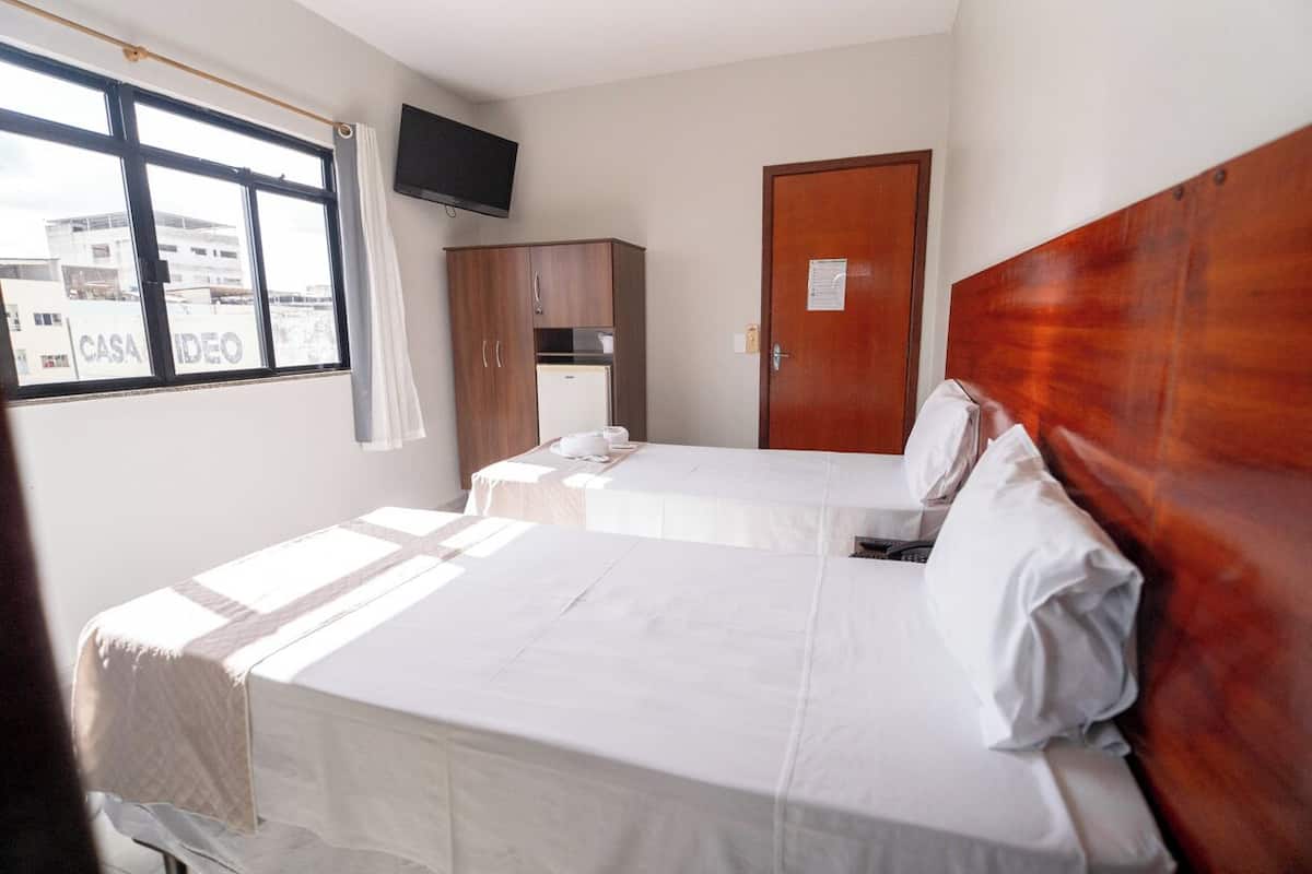 Quarto de hotel com duas camas, um guarda-roupa, televisão na parede e janela com vista para edifícios.
