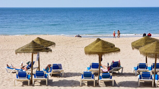 Een strand met rijen blauwe ligbedden en rieten parasols, waarvan sommige door mensen worden gebruikt.