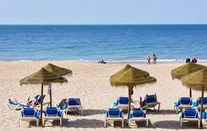 Una spiaggia con file di sedie a sdraio blu e ombrelloni di paglia, alcuni occupati da persone.