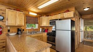 A wooden cabin kitchen with granite countertops, wooden cabinets, a stainless steel refrigerator, and a gas stove. - Three-Level Cabin with Mountain Views (Boone)