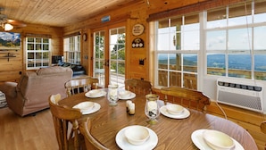 A wooden cabin interior with a dining table set for four, a sofa, and a view of the mountains through large windows. - Three-Level Cabin with Mountain Views (Boone)