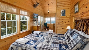 A wooden cabin bedroom with a large bed, a mounted deer head, and a view of the outdoors. - Three-Level Cabin with Mountain Views (Boone)