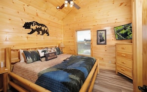 A cozy wooden cabin bedroom with a bed, a wooden dresser, a ceiling fan, and a bear-themed wall decal.