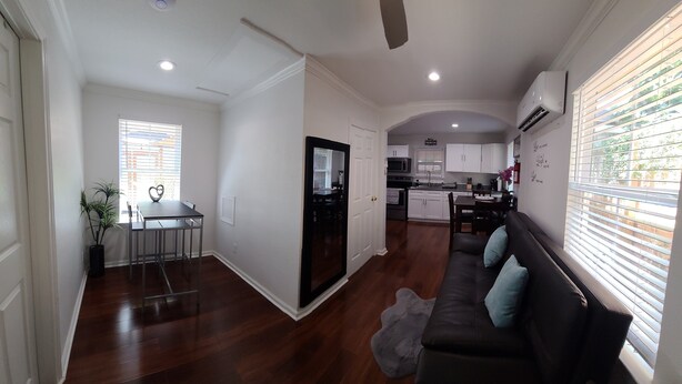 A spacious living area with a dark wood floor, a black sofa with blue cushions, a dining table with chairs, and a kitchen in the background.