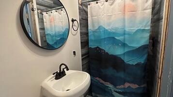 Une salle de bain avec un lavabo blanc, un miroir rond et un rideau de douche orné d’un motif représentant un paysage de montagne.