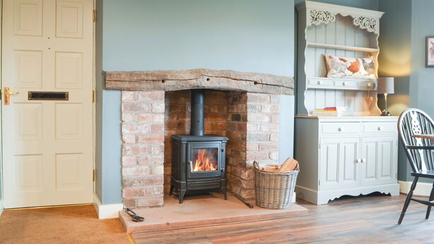 A room with a brick fireplace, a white cabinet with decorative pillows, and a wicker basket filled with firewood. - 3 bedroom accommodation in Westward, near Wigton (Westward, near Wigton)