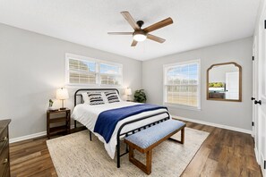Quarto com cama, mesas de cabeceira, ventilador de teto e janelas.