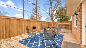 Eine Gartenpatio mit Holzdeck, einem blauen Teppich mit Muster, Metallstühlen und einem Grill.