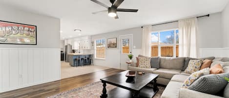 Un salon spacieux avec une table basse en bois foncé, un canapé beige agrémenté de coussins à motifs, un ventilateur de plafond et une grande fenêtre avec des rideaux.