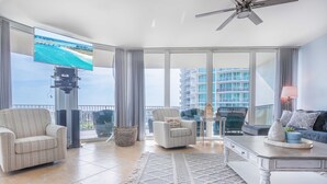 Ein geräumiges Wohnzimmer mit großem Fernseher, bequemen Sitzgelegenheiten und einem Balkon mit Blick auf das Meer und die Gebäude.