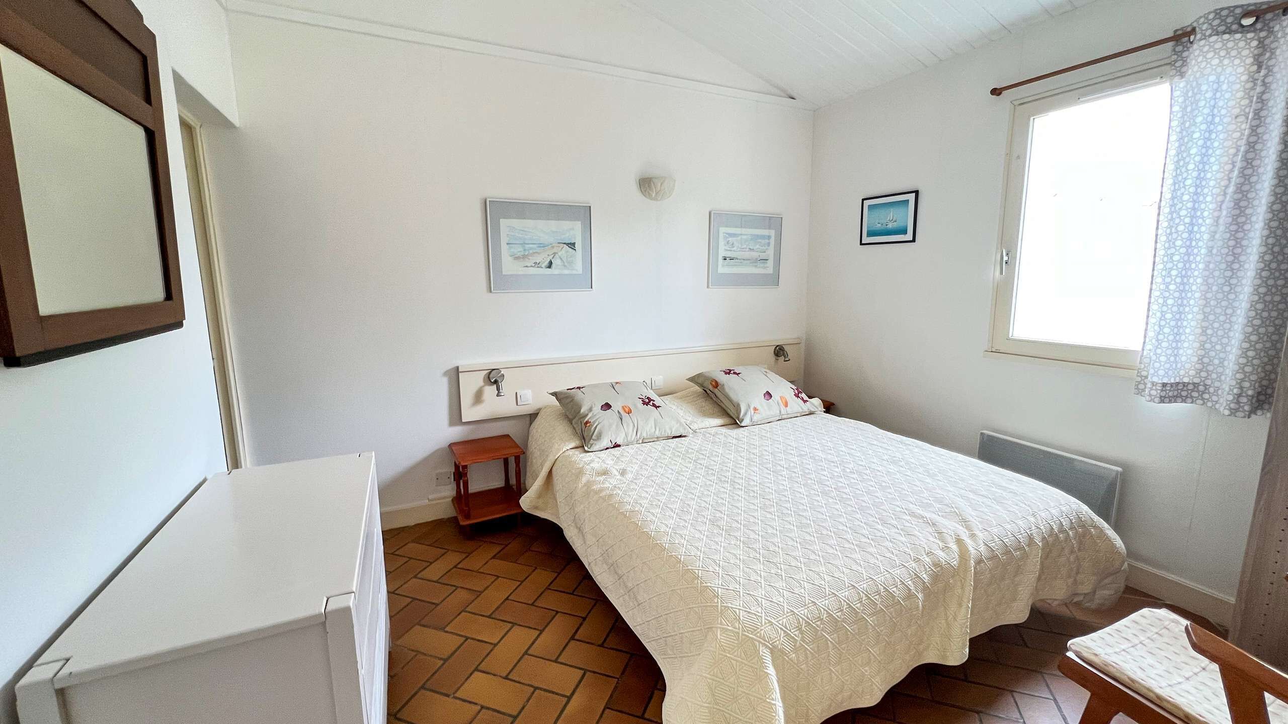 Een slaapkamer met een bed, een klein nachtkastje, een stoel, een raam met gordijn en twee foto's in een lijst aan de muur.