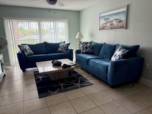 A living room with two blue sofas, a coffee table, a fan, and a painting on the wall.