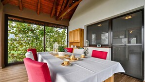 A dining area with a table set for four, red chairs, a wooden cabinet, and a large window with a view of greenery.