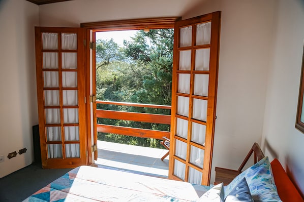 Een slaapkamer met een bed, een houten bankje en een balkon met uitzicht op bomen.