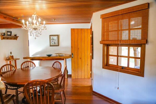 Un comedor con mesa y sillas de madera, una araña y un ventanal con persianas.