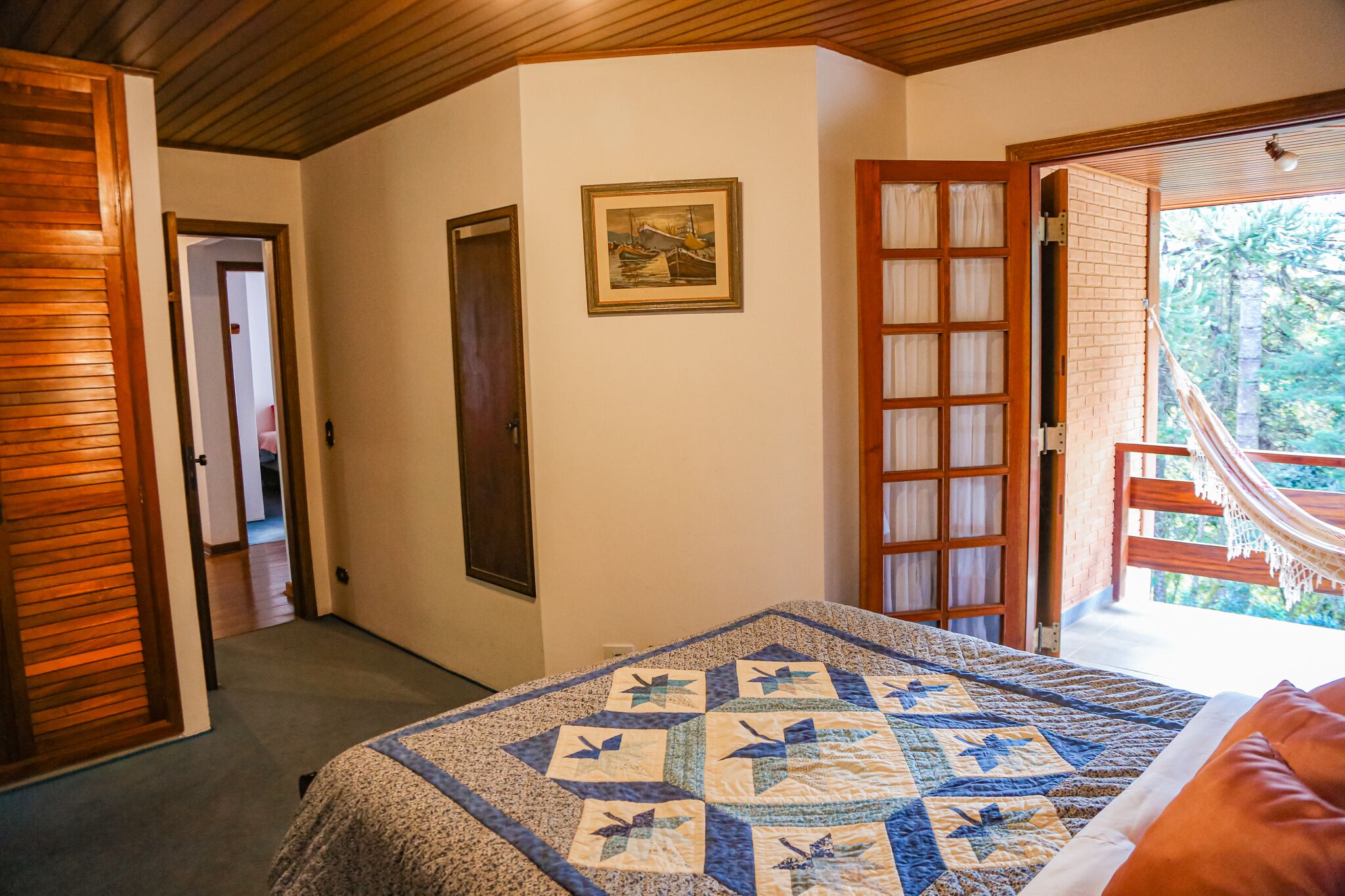A bedroom with a quilted bedspread, a door leading to another room, a framed picture on the wall, and a view of a hammock outside.