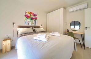 A modern bedroom with a large bed, a bedside table with towels, a desk with a chair, a wall-mounted air conditioner, and a colorful artwork above the bed. - Holiday apartment on the first floor with balcony (Njivice)