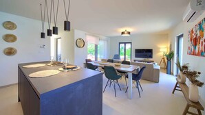 A modern kitchen with a central island, dining area with chairs, a sofa, and a TV in the background.