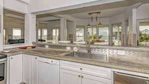 A modern kitchen with white cabinetry, a large island with a sink, and a view of the outside through large windows. - Ocean front home, access to both clubs and large porches! Breaking Point (Bald Head Island)
