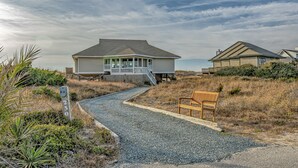 A coastal house with a gravel driveway leading to a wooden bench. - Ocean Front and beach access to West Bald Head at the Wetmore House! Pet Friendly! (Bald Head Island)