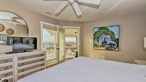 A bedroom with a bed, a TV, a dresser, a ceiling fan, a painting, and a door leading to a balcony. - Ocean Front and beach access to West Bald Head at the Wetmore House! Pet Friendly! (Bald Head Island)