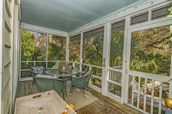 A covered outdoor area with a table and chairs, surrounded by a white railing, overlooking a garden with trees and plants.