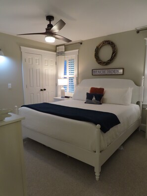 A bedroom with a bed, a ceiling fan, a window with blinds, a bedside table, and a wall sign that reads 'BEACH HOUSE'.