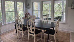 A dining area with a large table and chairs, large windows with a view of greenery, and a sign above the table reading "EAT, DRINK, AND BE MERRY".