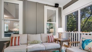 Ein gemütliches Wohnzimmer mit einer Couch, Sesseln und einem Couchtisch. Große Fenster bieten Blick auf Bäume und es gibt eine weiße Geländer.