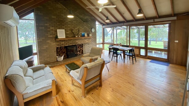 A spacious living room with a fireplace, wooden furniture, and large windows offering a view of the outdoors. - Charming off-grid cabin with room to explore (Briagolong)