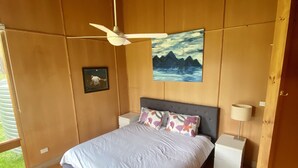 A bedroom with a bed, wooden walls, a ceiling fan, a painting, and a nightstand with a lamp. - Charming off-grid cabin with room to explore (Briagolong)