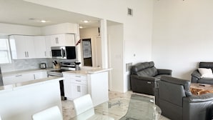 Een moderne keuken met witte kasten, een eethoek met een glazen tafel en stoelen, en een zithoek met een leren bank en een salontafel.