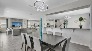 A modern kitchen with white cabinets, a central island, and a dining area with a dark table and grey chairs. - 5 Star Villa Monarchia – A Realm of Refined Power (Orlando)