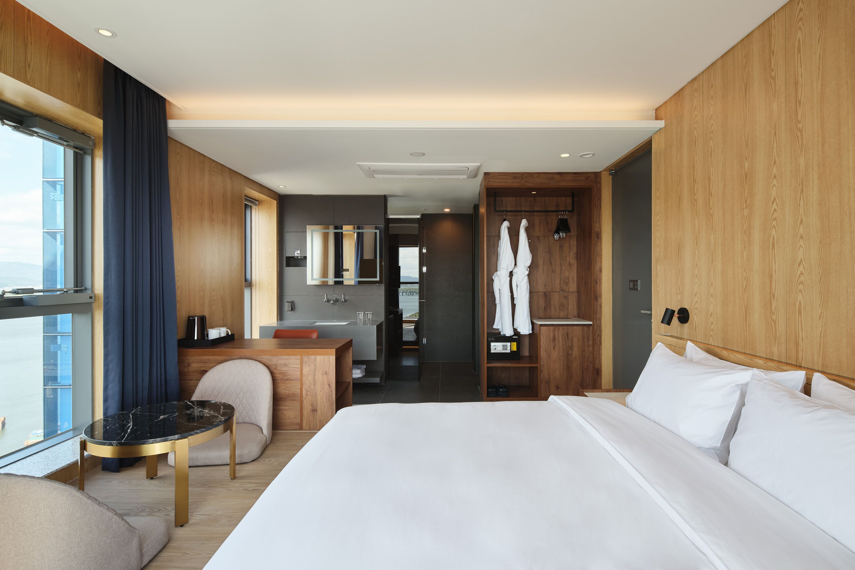 A modern hotel room with a large bed, a small table, a seating area, a bathroom, and a view of the city.