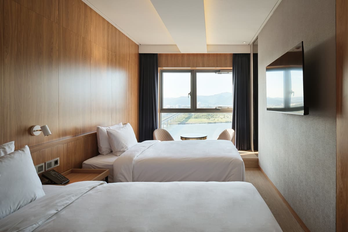 A hotel room with two beds, a TV, a window with a view of a bridge and landscape, and a wooden wall.