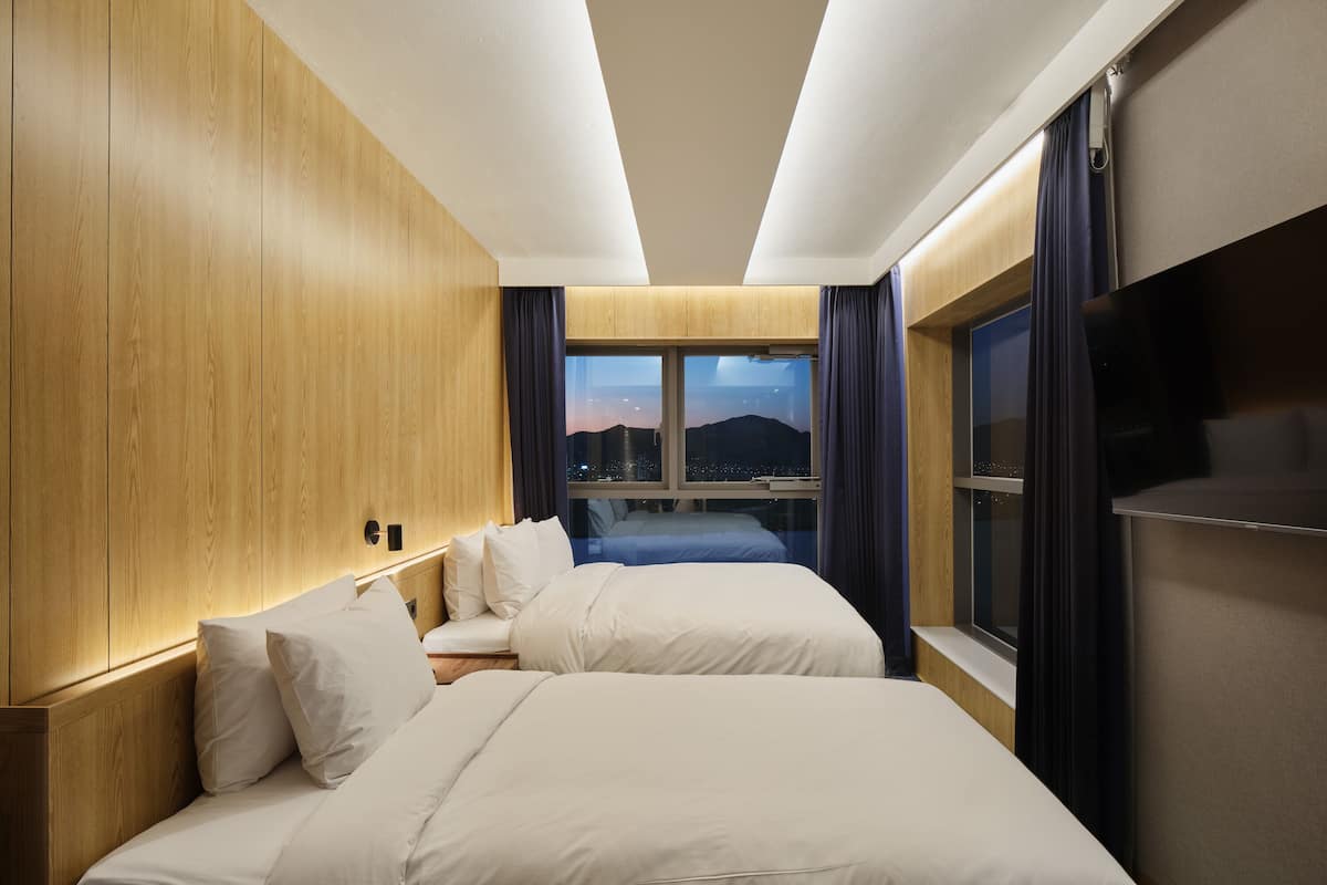 A hotel room with two beds, wooden paneling, a large window with a city view, and a flat-screen TV.