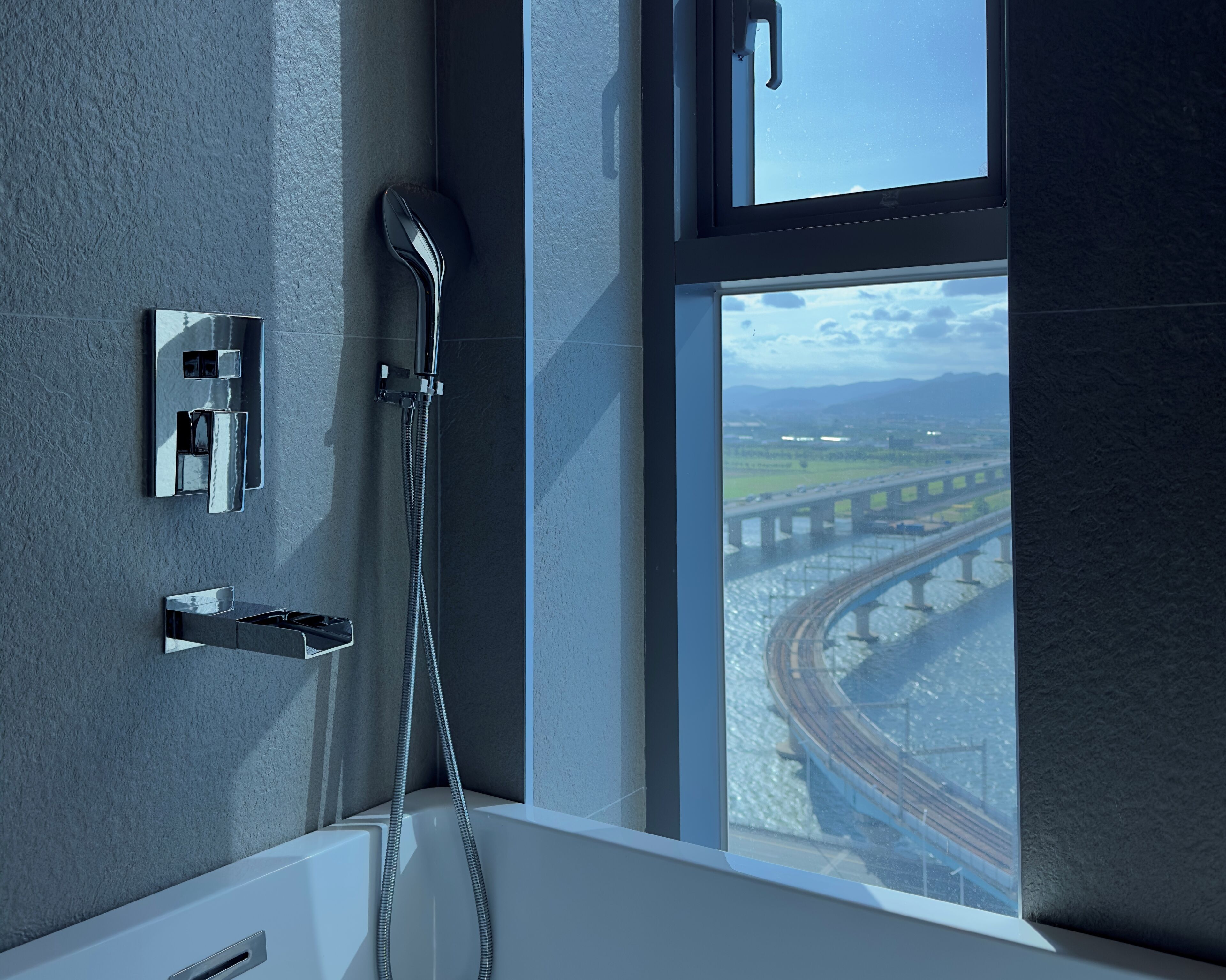 A modern bathroom with a bathtub, a handheld showerhead, and a window overlooking a bridge and railway tracks.