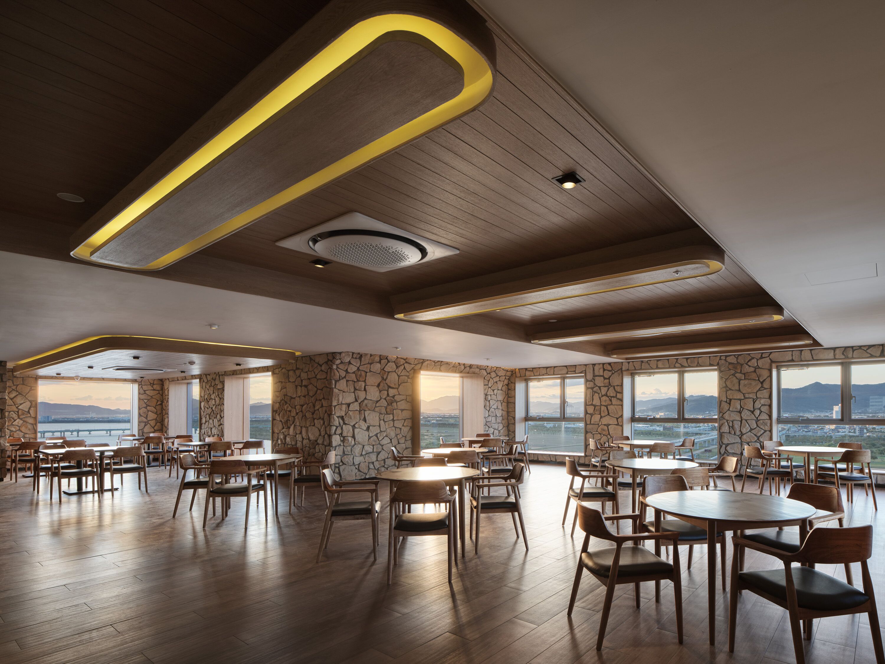 A spacious dining area with wooden flooring and stone walls, featuring multiple tables and chairs, large windows with a view of the mountains.