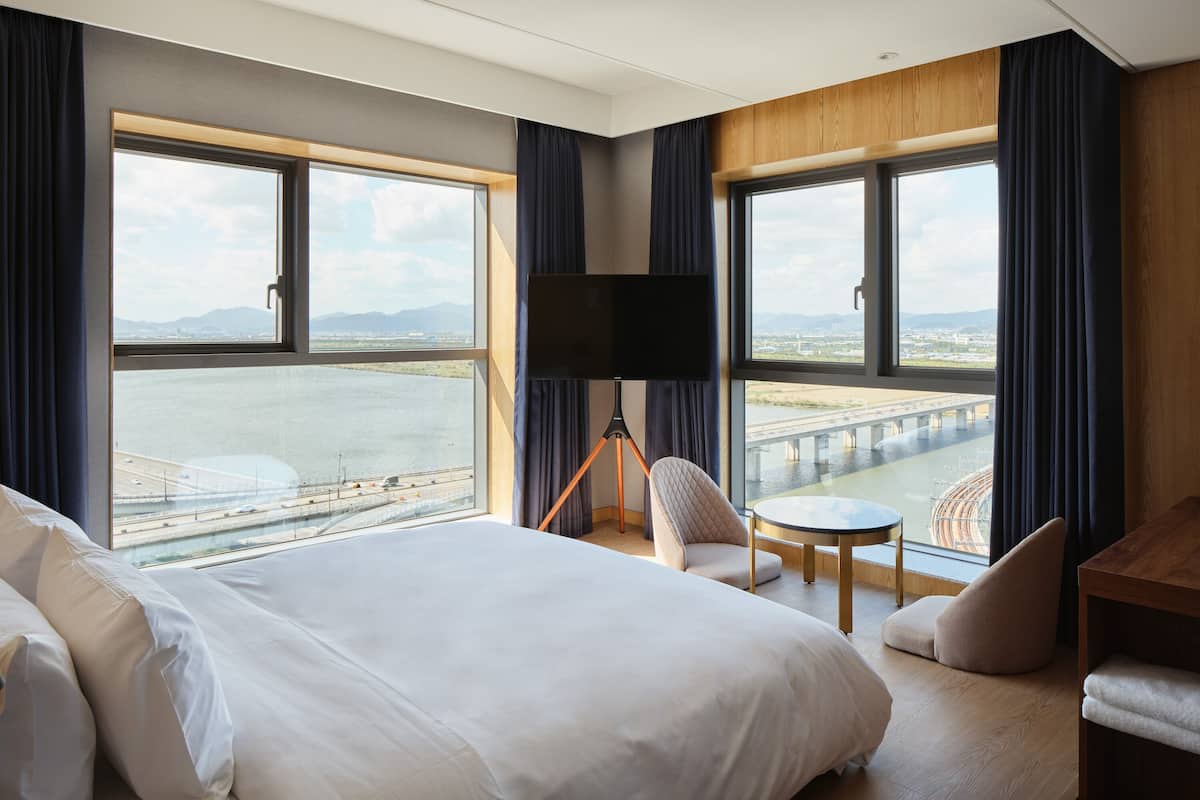 A hotel room with a large bed, a desk, a chair, and a TV. There are large windows with a view of a bridge and water.