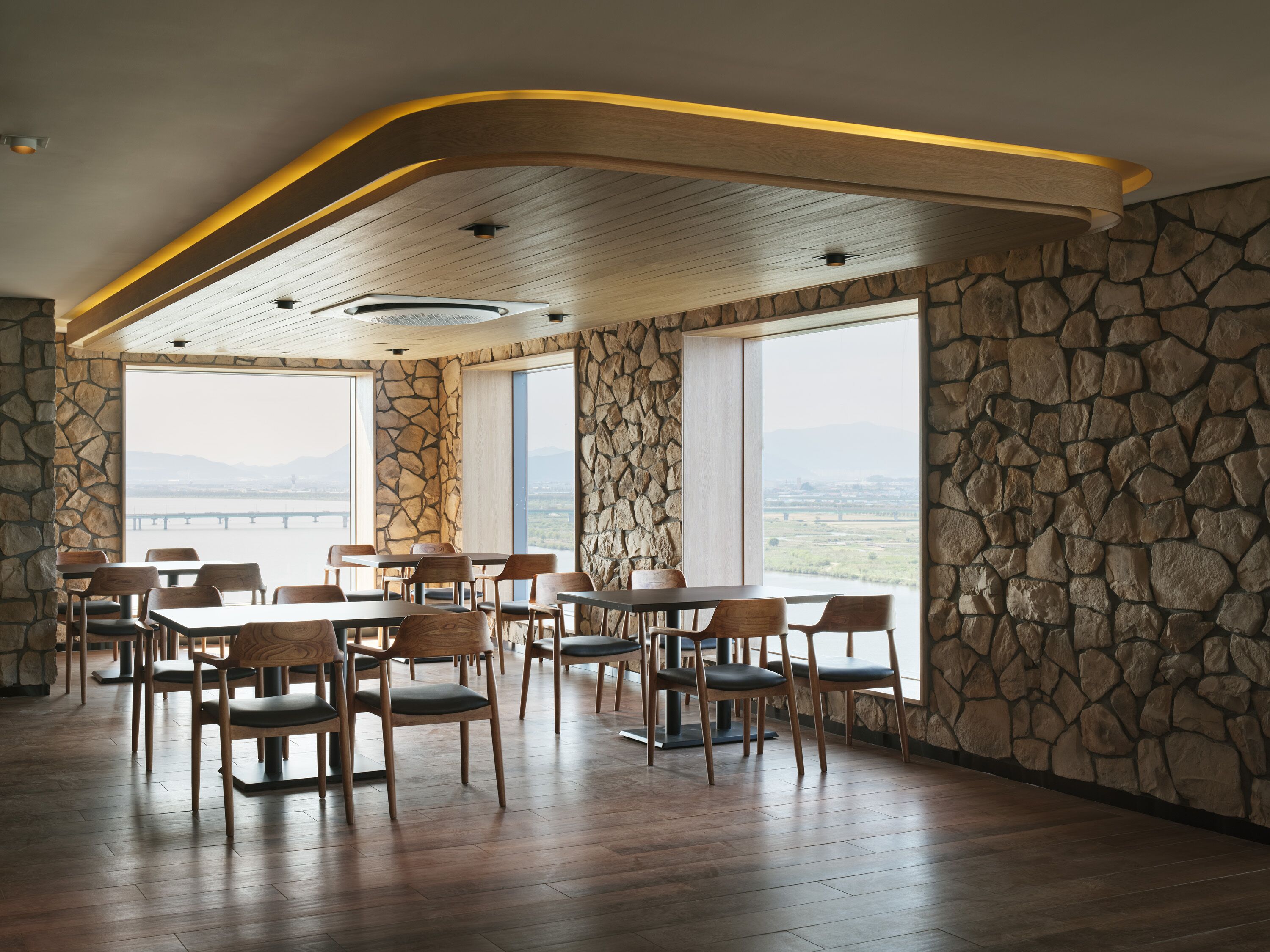 A restaurant with stone walls, wooden tables, and chairs, large windows with a view of a bridge and mountains.