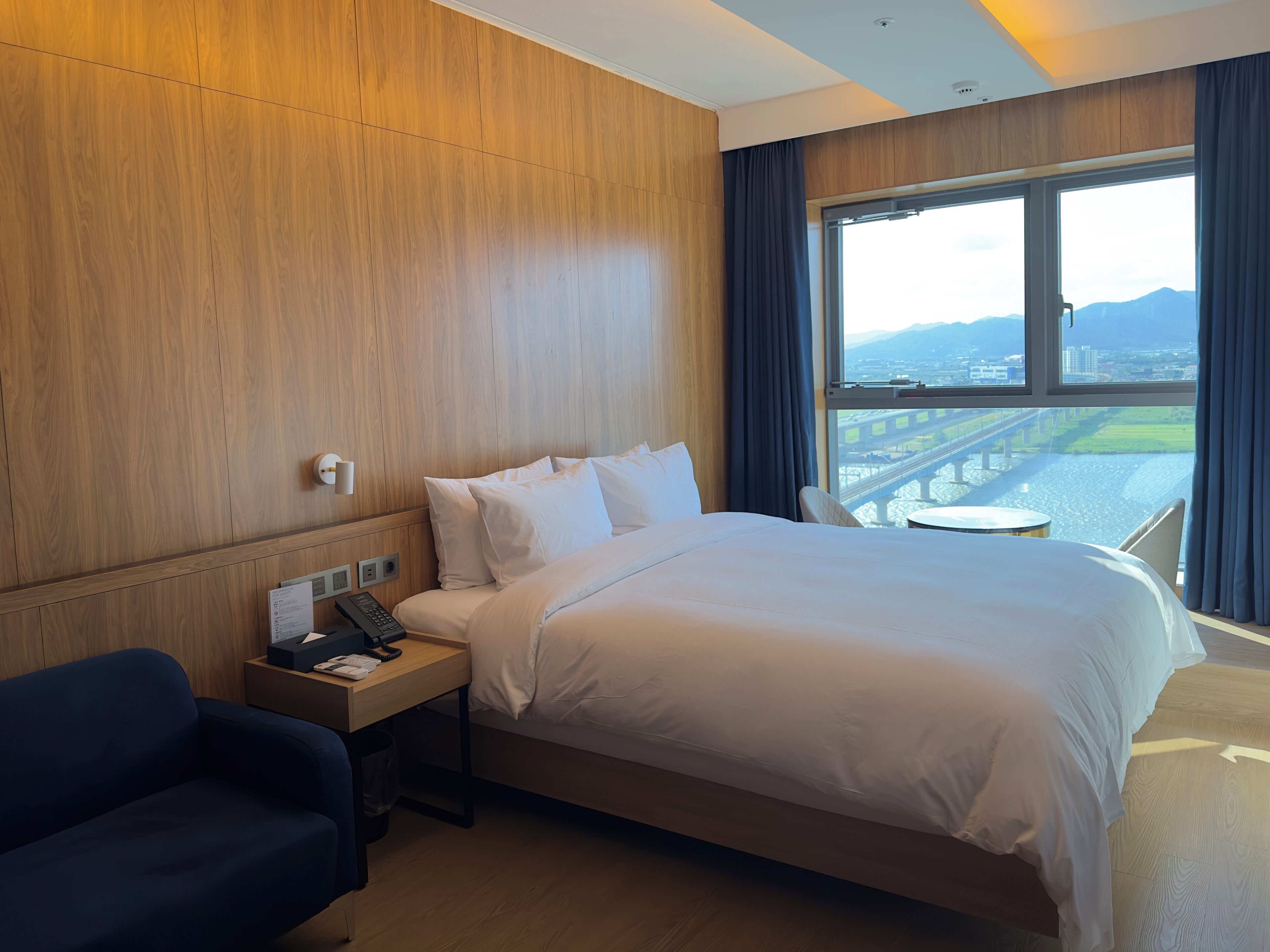 A hotel room with a large bed, a desk with a phone and papers, a chair, and a view of a bridge and cityscape through a large window.