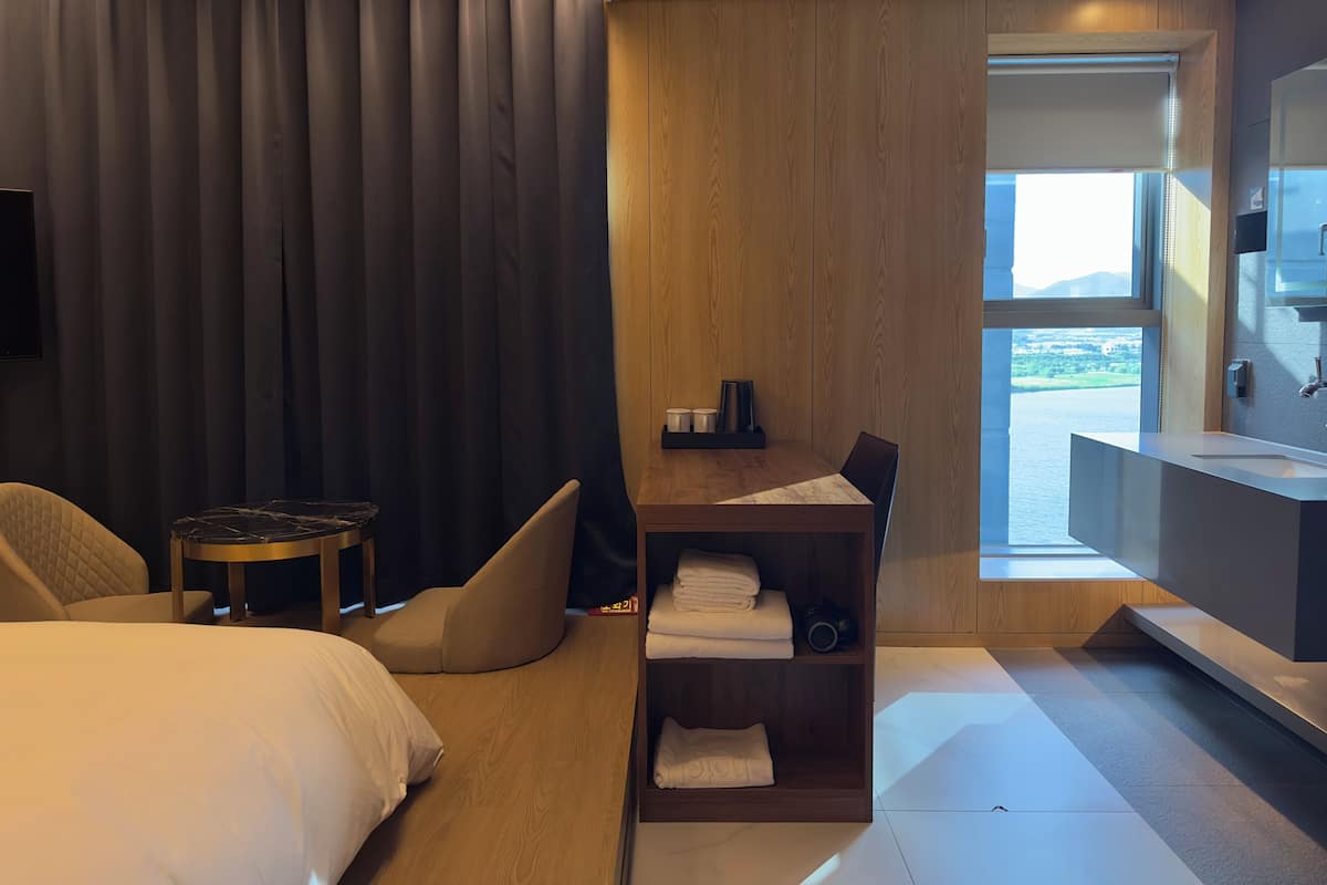A modern hotel room with a large bed, a small desk, a seating area, and a view of a body of water through a large window.