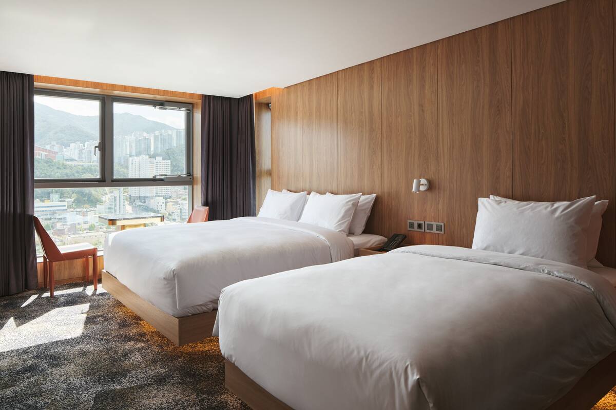 A hotel room with two beds, a wooden headboard, a city view, and a small table.
