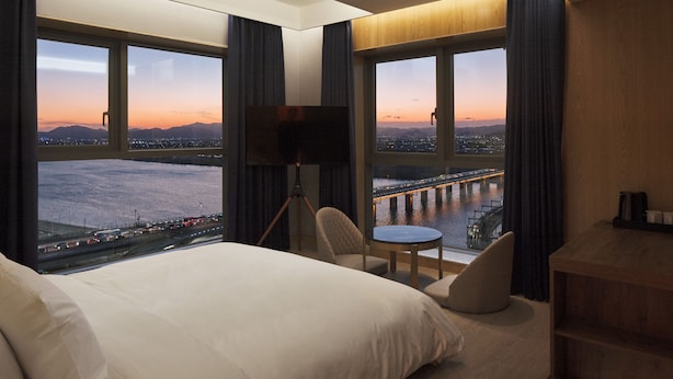 A hotel room with a large bed, a desk, a chair, and a TV. There are large windows with a view of a city and a bridge. - hotel tt Gupo (Busan)