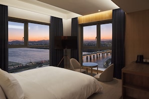 A hotel room with a large bed, a desk, a chair, and a TV. There are large windows with a view of a city and a bridge. - hotel tt Gupo (Busan)